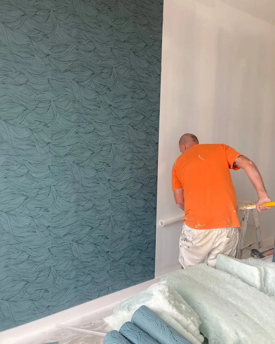 Jef in an orange t-shirt hanging teal wave-patterned wallpaper